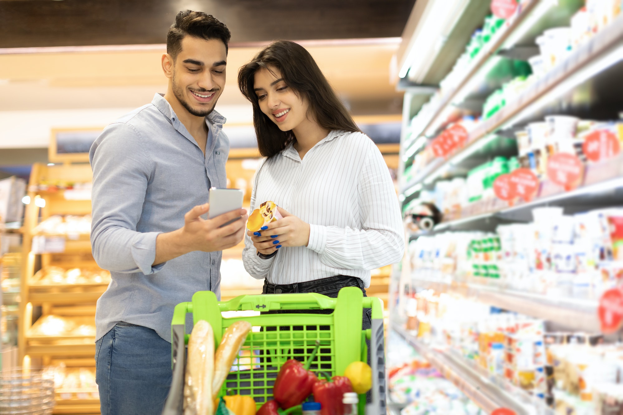 Arab Couple Using Smartphone Buying Food Shopping In Modern Supermarket